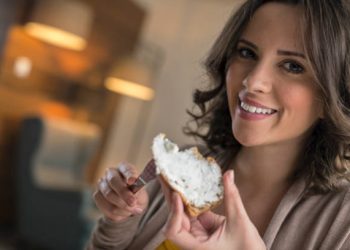 Young women spreading cream cheese on slice of bread and eating at home